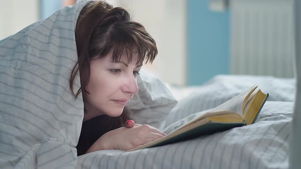Pretty Brunette Woman Lies in Bed and Reads a Book in the Morning
