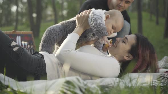 Carefree Smiling Beautiful Mother Lying on Picnic with Infant As Little Son Feeding Father Croissant alt