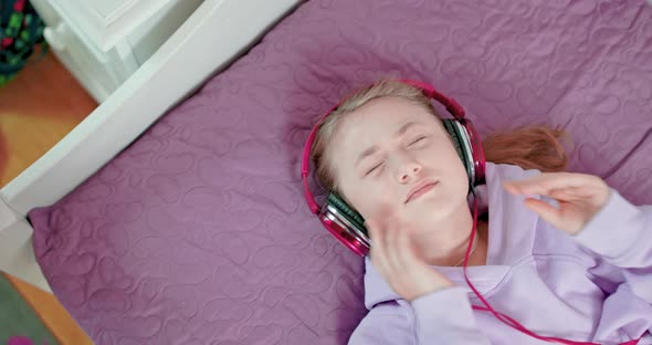 Teen Girl is Singing with Closed Eyes Lying on Her Bed in Bedroom Wearing Headphones alt