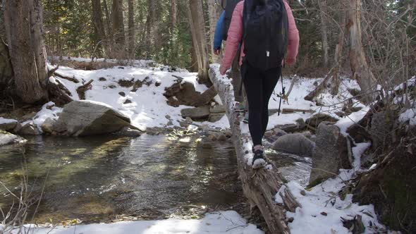 Couple hiking on log covered in snow over river as they balance alt