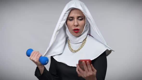 Absorbed Caucasian Woman in Nun Costume Lifting Dumbbell Watching Online Lesson in Smartphone alt