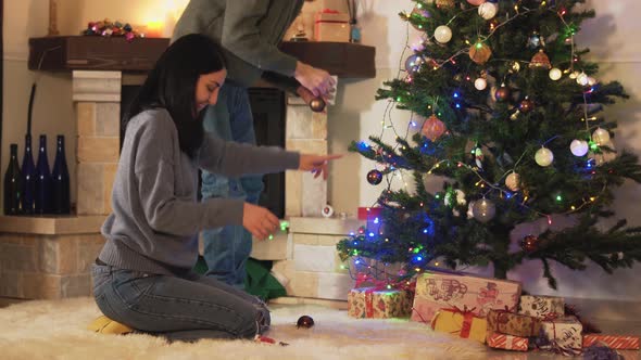 Man and Woman Decorating Christmas Tree in Modern Room. Girl Gives Toys, Man Hanging Them on alt