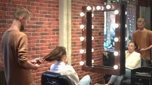 Side View Young Male Hairstylist Examining Long Hair of Female Client Sitting in Armchair in Beauty alt