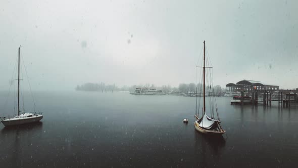 Snow falling over Maggiore lake and anchored sailing boat and ferry, Italy alt