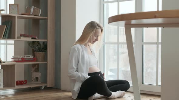 Happy pregnant woman is sitting on the floor of room near large window