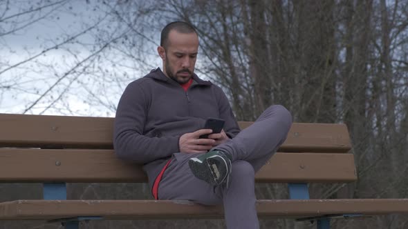 Closer shot of young bearded man with short hair sitting on a park bench on a cloudy fall day and pl alt