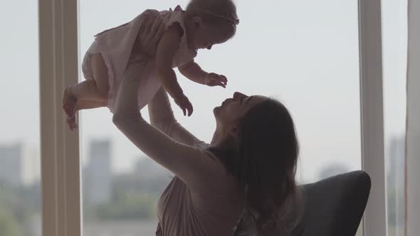 Cute Young Woman Sitting in the Comfortable Chair in Front of the Window Holding Baby Girl in Her alt
