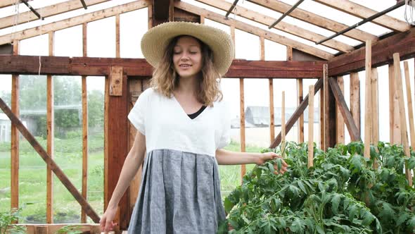 Girl Farmer in a Straw Hat and Dress Goes To the Greenhouse and Enjoys the Harvest alt