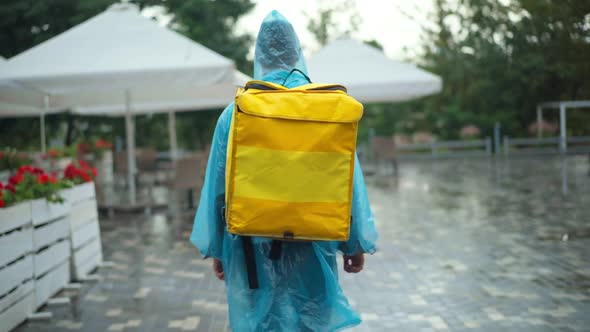 Back View Courier in Rain Coat and Coronavirus Face Mask with Backpack Walking on City Street alt