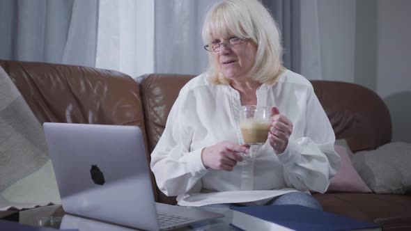 Intelligent Senior Woman in Eyeglasses Watching Pictures or Photos on Laptop Screen and Drinking alt