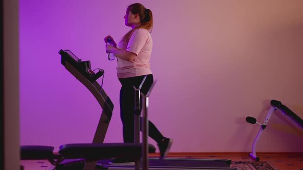 Side View of Plump Caucasian Woman Walking on Treadmill Drinking Refreshing Water alt