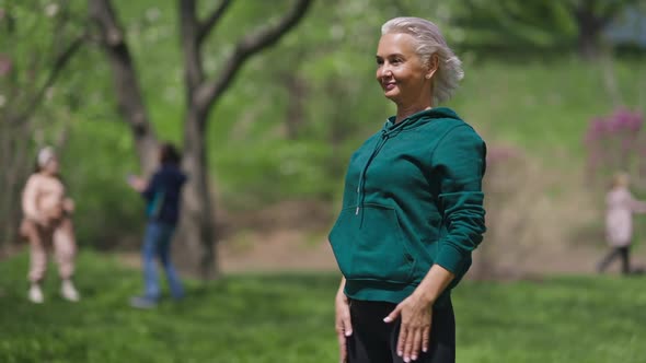 Positive Slim Female Retiree Bending Forward Smiling Working Out in Sunny Summer Spring Park alt