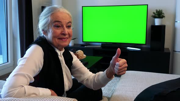 Old Caucasian Woman Watches Television in Living Room and Then Shows Thumb Up alt