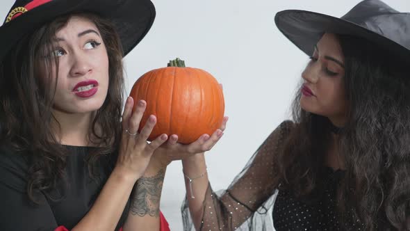 Two Colombian Girl Friends In Witch Halloween Costumes Both Holding A Pumpkin Isolated On White Wall alt