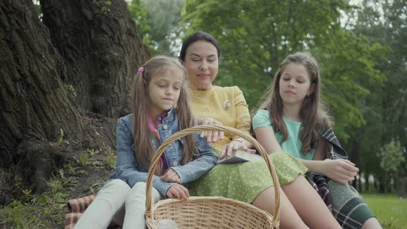 Mature Woman Sitting on the Grass Under the Tree in the Park with Two Cute Granddaughters alt