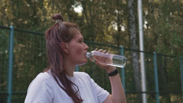 Woman Drinks Water From a Bottle While Sitting on Sports Platform After Training