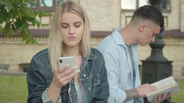 Portrait of Pretty Blond Girl Listening To Music in Earphones with Blurred Concentrated Boy Reading alt