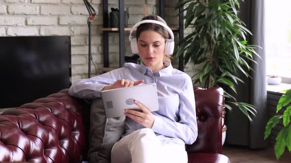 Young attractive smiling caucasian woman using tablet sitting on sofa at home, singing song, enjoyin alt