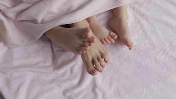 Two Pairs of Legs of the Family in Bed Under the Coverlet - Mother and Baby alt