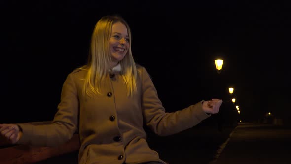 A Young Woman Sits on a Bench and Dances While Smiling at the Camera in an Urban Area at Night alt