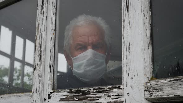 Man In Medical Mask Looks Sadly Out Of Window At Street Because Of The Pandemic alt