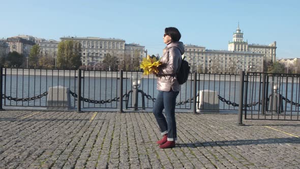 A Middleaged Woman Brunette with a Bouquet of Yellow Leaves Walks Along the City Embankment alt