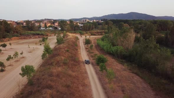 Aerial done shot flying forward of a grey car driving on a gravel road with a town at the background alt