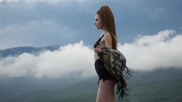 Bright Young Brunette Girl in Short Shorts and Cape Stands High in the Mountains