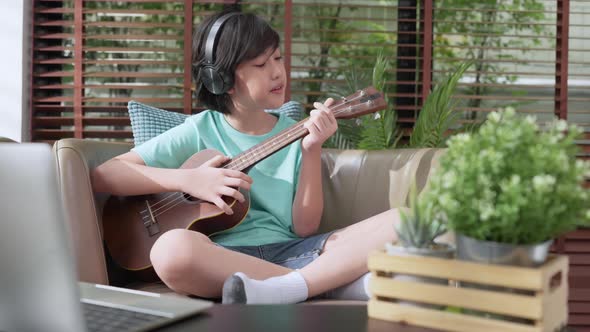 Little asian child boy online learning playing ukulele on armchair at living room