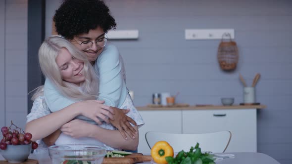 Young Girl Hugging and Kissing Her Girlfriend From the Back Cutting Vegetables