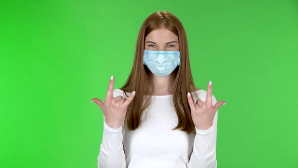 Portrait of Pretty Young Girl in Medical Protective Face Mask Looking at Camera and Making a Rock alt