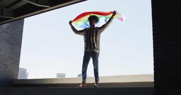 African american man holding lbgt flag while standing near the window alt