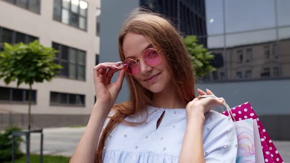 Young Woman in Pink Sunglasses Standing in Front of the Shopping Mall and Holding Shopping Bags alt