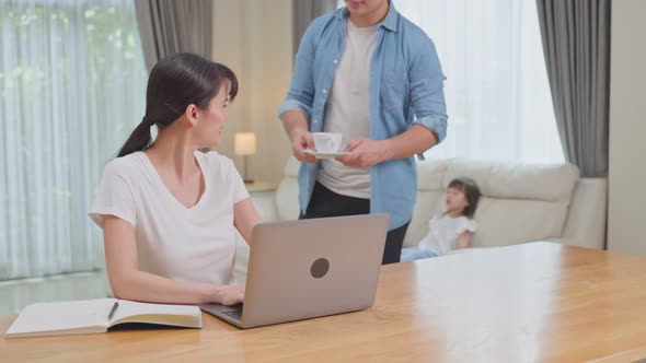 Asian loving husband take care of beautiful freelance worker wife by serve a cup of coffee on table alt