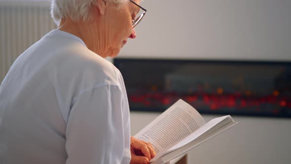 Aged lady with short grey hair and glasses reads book sitting on sofa alt