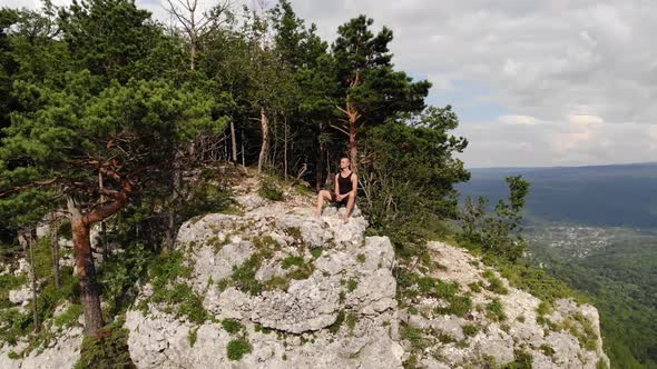 A Long-haired, Thin Man in a Black T-shirt and Shorts Sits on the Edge of a Cliff and Looks Out at alt