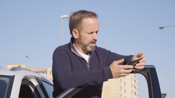 A Middleaged Handsome Caucasian Man Scrolls on a Smartphone As He Stands Next to His Car