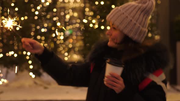 Young Smiling Woman Holding Burning Sparkler and Coffee in Cold Winter Evening on Street
