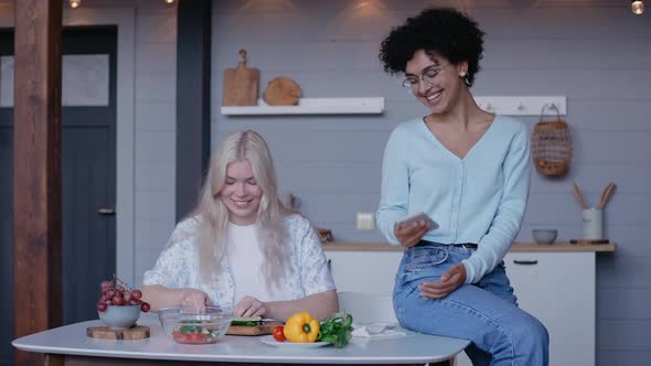 Young Girl Cuts Vegetables and Laughs with Her Girlfriend Sitting on a Table