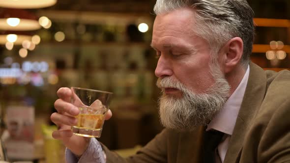 Close-up of a gray-haired bearded man enjoying whiskey alone in a bar alt