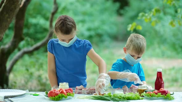 Little boy and girl in medical masks sprinkle spices on raw meat. Evening picnic in nature