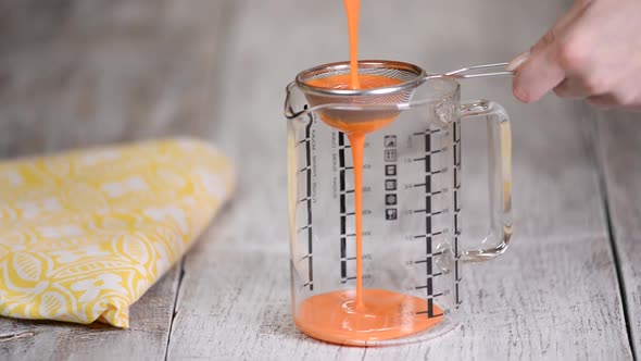 Woman sieve the mix of mirror glaze into a large jug to remove any excess bubbles. alt