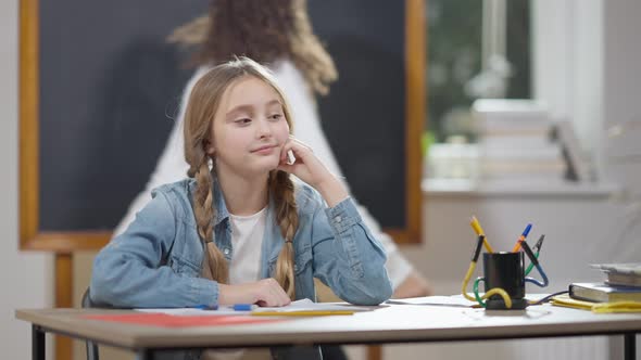 Bored Sad Caucasian Schoolgirl Sitting at Desk in Classroom with Blurred African American Classmate alt