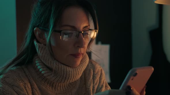 Pretty woman in eyeglasses late at night scrolling with smartphone