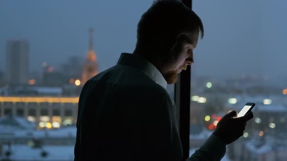 a man uses the phone by the window overlooking the city at night