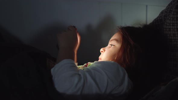 Baby Girl is Watching Video on Smartphone in Bed Before Sleeping at Evening alt