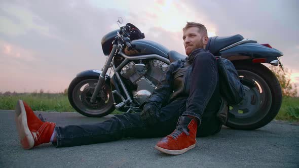 Middleaged Biker with Beard Sits By Motorcycle at Sunset