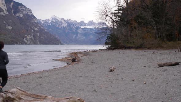 Sporty couple running on the stony shore of a lake in front of a mountain range with snowy peaks alt