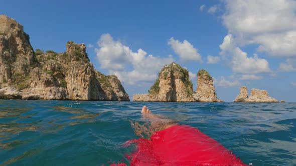 Fpv of male legs and feet relaxing while floating on sea water with Scopello Stacks or Faraglioni in alt