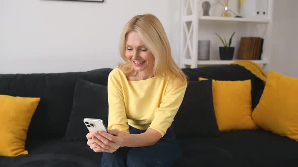 Serene 50s Middleage Woman Using Smartphone Sitting on the Sofa at Home